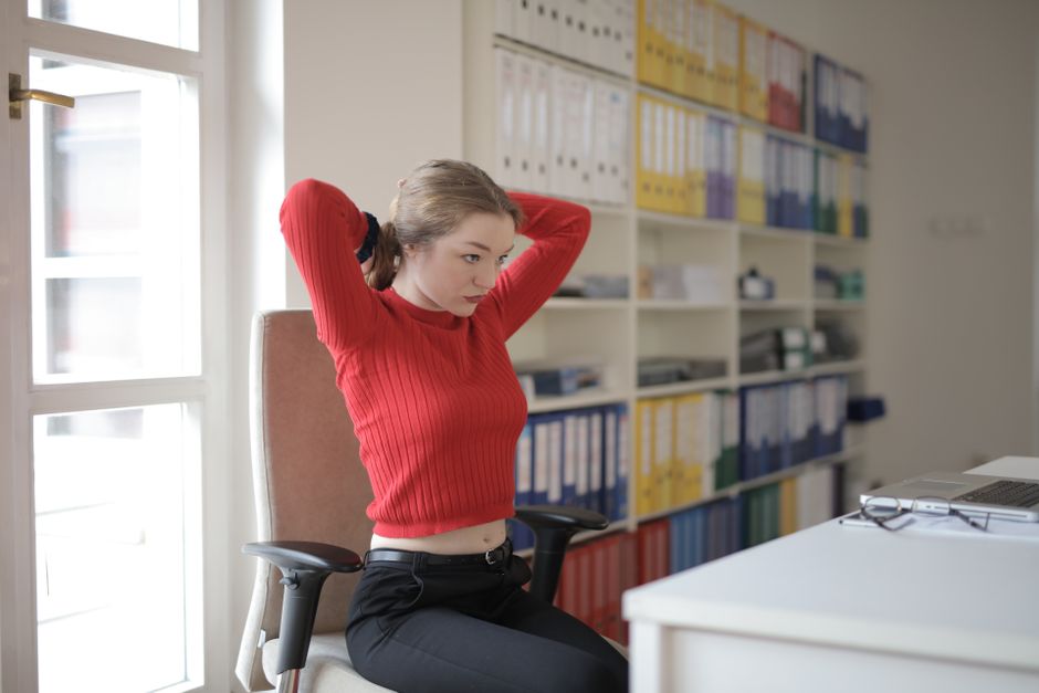 An office worker standing beside their desk, performing a full-body back stretch with arms extended overhead, illustrating a break from sitting.