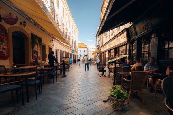 A charming, narrow European street scene with outdoor cafes, local residents enjoying coffee, and a tram passing by, emphasizing local life and transit.