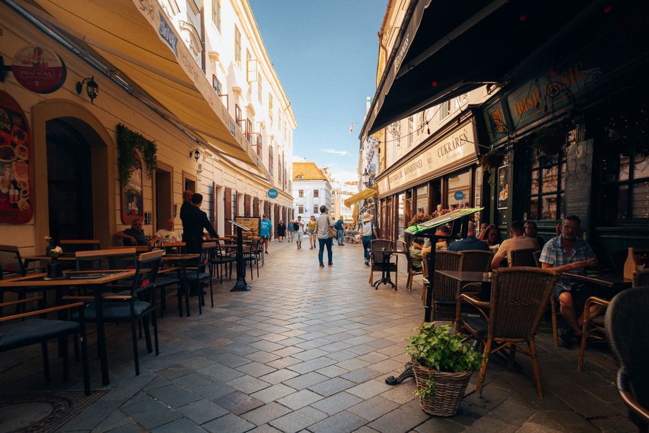 A charming, narrow European street scene with outdoor cafes, local residents enjoying coffee, and a tram passing by, emphasizing local life and transit.