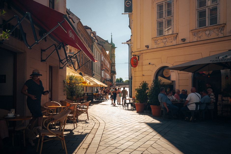 An aerial view of a charming outdoor cafe with red umbrellas, tables spilling onto a cobblestone street in a historic European city, passersby enjoying a leisurely pace.