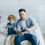 A parent and child sharing a controller, playing a video game on a console, illustrating shared gaming experiences.