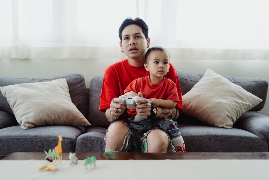 A parent sitting next to a child, patiently showing them how to use a game controller while looking at a console screen.