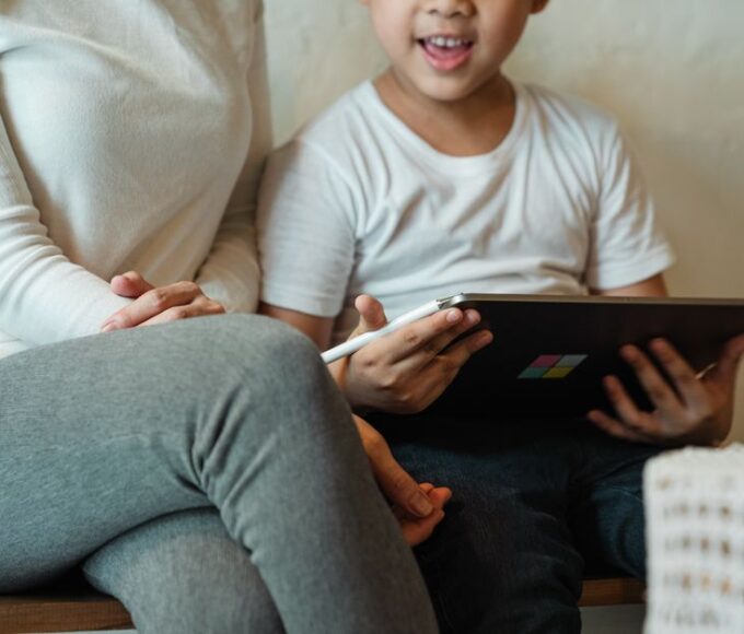 A parent and child are sitting together, looking at a tablet screen displaying Roblox, with the parent pointing at the screen, symbolizing shared understanding of safety settings.