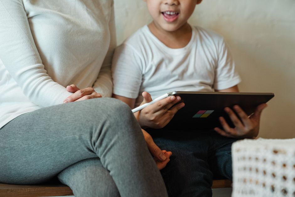 A parent and child are sitting together, looking at a tablet screen displaying Roblox, with the parent pointing at the screen, symbolizing shared understanding of safety settings.