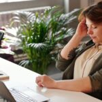 A parent with visible neck strain working at a cluttered home desk, laptop screen too low, highlighting common ergonomic mistakes.