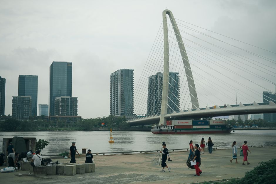 A wide, modern riverfront promenade with people walking, jogging, and cycling, featuring contemporary architecture and a scenic bridge in the background.