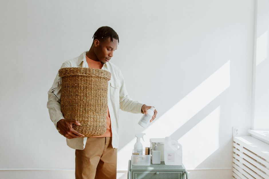 A person wearing a simple apron, cleaning a kitchen counter with a spray bottle and a cloth, with blurred green plants in the background, suggesting natural cleaning.