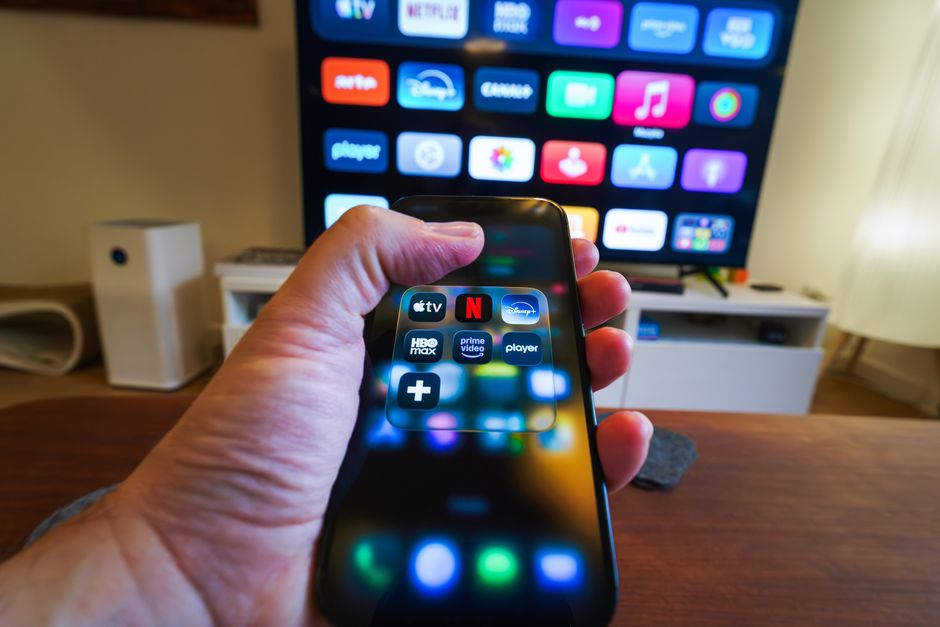 A person's hands holding a smartphone displaying various gaming subscription logos side-by-side, such as Xbox Game Pass, PlayStation Plus, and Nintendo Switch Online, illustrating a comparison of services.