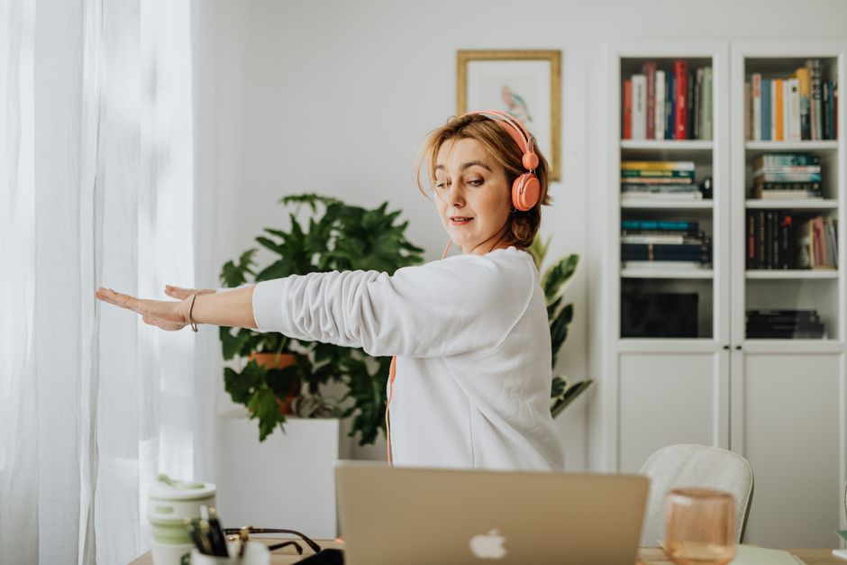 A person with a neutral expression performing a gentle neck rotation stretch while sitting at an ergonomic desk, in a bright office environment.