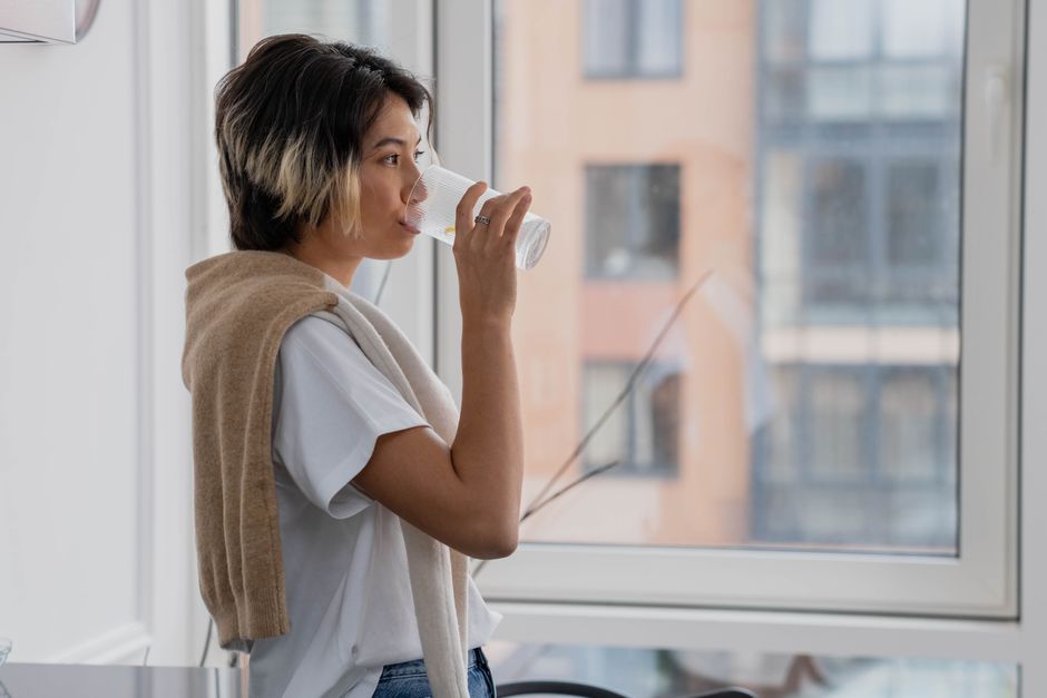 A remote worker sitting at a desk in a warm, urban apartment, taking a mindful sip from a reusable water bottle, with a fan in the background.