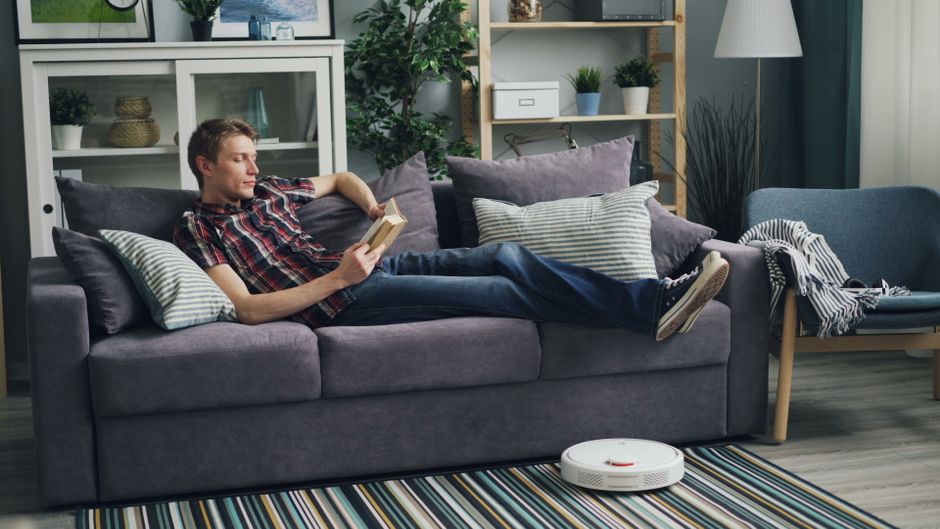 A person relaxing on a sofa with a book, while a robot vacuum cleans in the background, representing the benefits of automating mundane tasks.