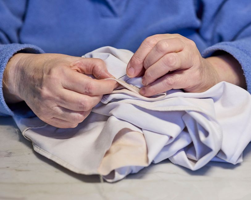 Close-up shot of hands carefully stitching a small tear in a blue cotton shirt. A needle and thread are visible, demonstrating the act of mending.