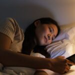 A person's hand holding a smartphone displaying a sleep tracking app's detailed graphs and data, with a blurred bedroom in the background, illustrating data interpretation.