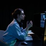 A person sitting at a desk, looking intently at two computer monitors, one displaying work documents and the other a game, illustrating the dual purpose.