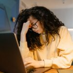 A person with glasses rubbing their eyes in front of a laptop, looking tired and overwhelmed by digital screens.