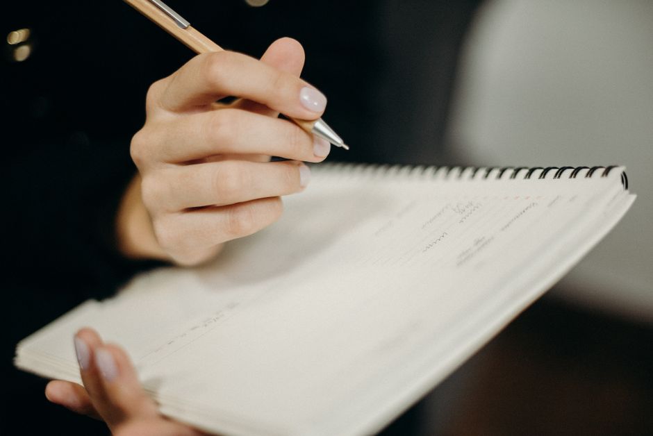 A hand holds a pen, poised over a notebook with a detailed shopping list written down, next to a smartphone showing online shopping tabs.