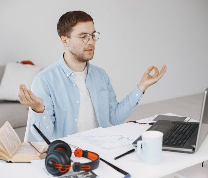 A person sitting calmly at a home office desk, eyes closed, possibly meditating, with a laptop in the background, symbolizing peace amidst remote work.