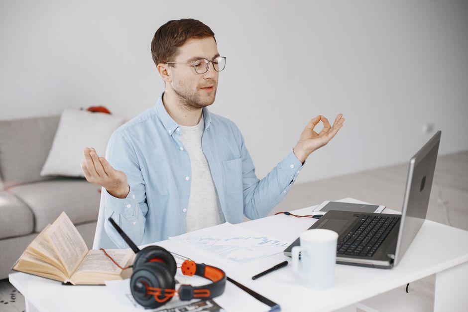 A person sitting calmly at a home office desk, eyes closed, possibly meditating, with a laptop in the background, symbolizing peace amidst remote work.