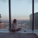 A person meditating calmly in a sunlit city apartment, overlooking urban buildings, embodying a slow morning routine.
