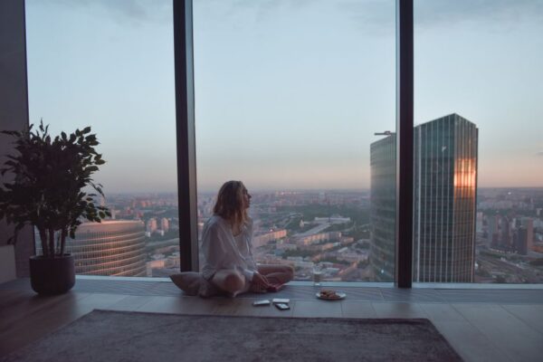 A person meditating calmly in a sunlit city apartment, overlooking urban buildings, embodying a slow morning routine.