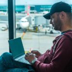 A person using a laptop with world map overlay in a modern airport lounge, depicting international work and travel.