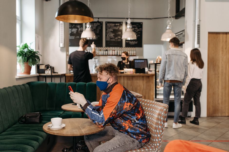 A person is focused on their smartphone in a bustling cafe, with Wi-Fi signals indicating public network use, emphasizing the need for VPN security in such environments.