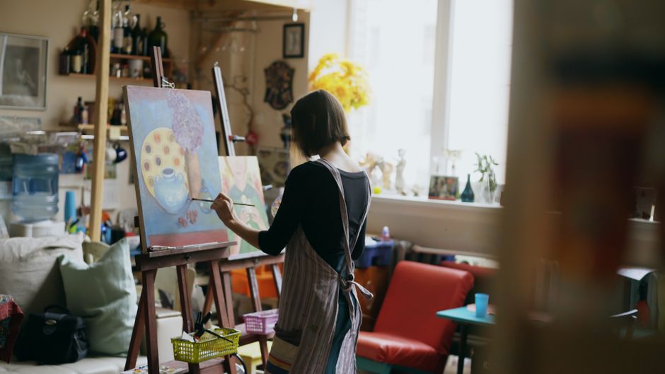 A person focused on painting on a canvas in a sunlit home studio, surrounded by art supplies.