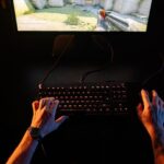 A close-up shot of hands on a keyboard and mouse, with a complex turn-based strategy game displayed on a computer monitor in the background.