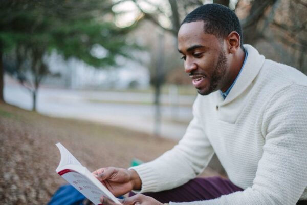 A person relaxing on a park bench, reading a book, with trees and city buildings in the background.