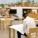 A person comfortably reading a book in a modern, well-lit public library, surrounded by shelves and other patrons using computers.