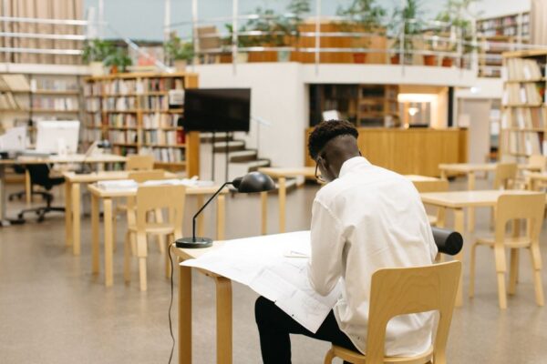 A person comfortably reading a book in a modern, well-lit public library, surrounded by shelves and other patrons using computers.