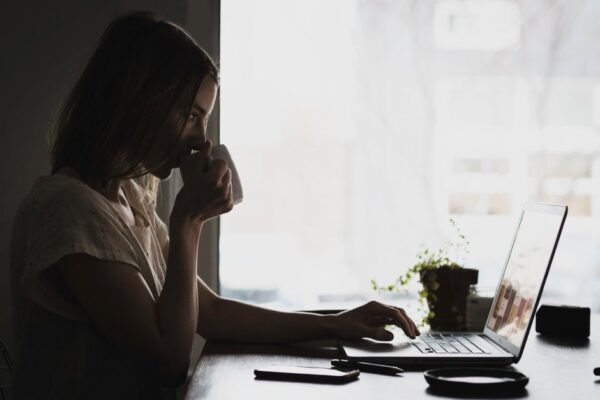 A person in a business casual shirt sips coffee from a mug while looking at a laptop screen, with soft morning light filtering in.