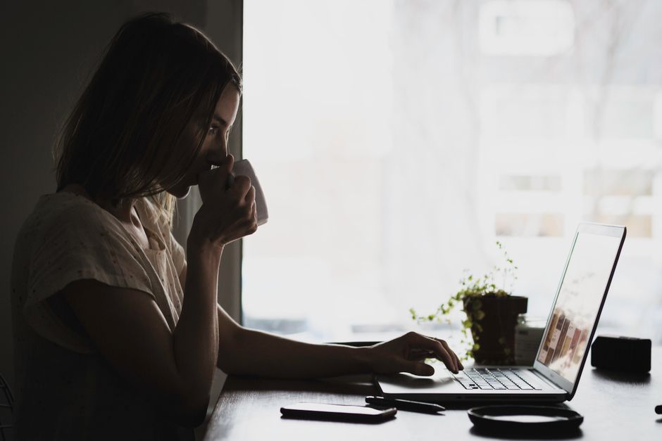 A person in a business casual shirt sips coffee from a mug while looking at a laptop screen, with soft morning light filtering in.