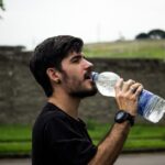 Person sipping from a reusable water bottle during a hot summer day, with sweat on their brow.