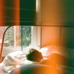 A person peacefully sleeping in a bed in a modern apartment, subtly showing earplugs, with a blurred city skyline visible through a window in the background.
