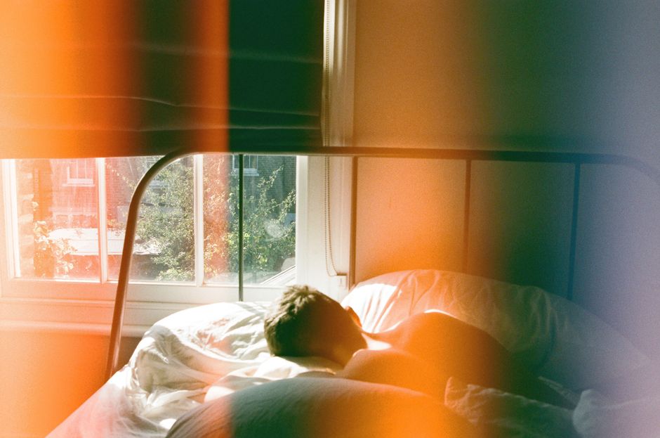 A person peacefully sleeping in a bed in a modern apartment, subtly showing earplugs, with a blurred city skyline visible through a window in the background.