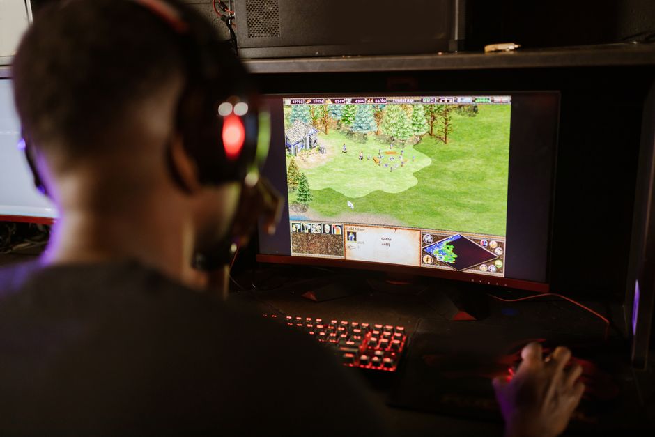 A close-up of hands on a keyboard and mouse, navigating a strategy game interface displaying ancient historical architecture and military units.