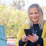 A person smiling while using a sleek, modern budget smartphone outdoors, clearly enjoying the experience.