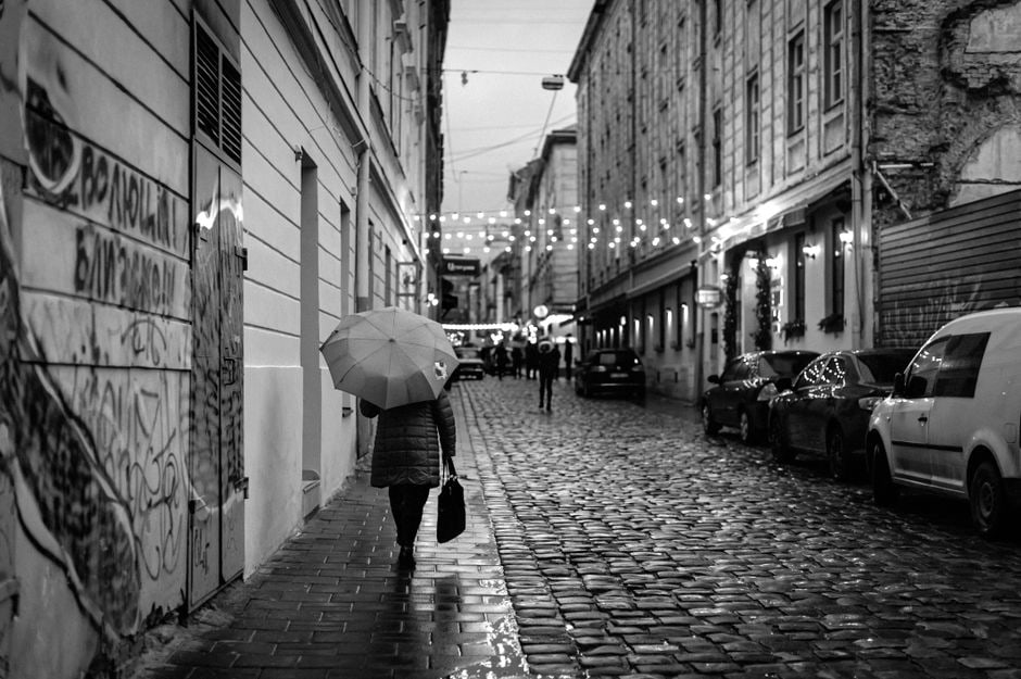 A person with a colorful umbrella walking on a wet, historic cobblestone street, puddles reflecting city lights.