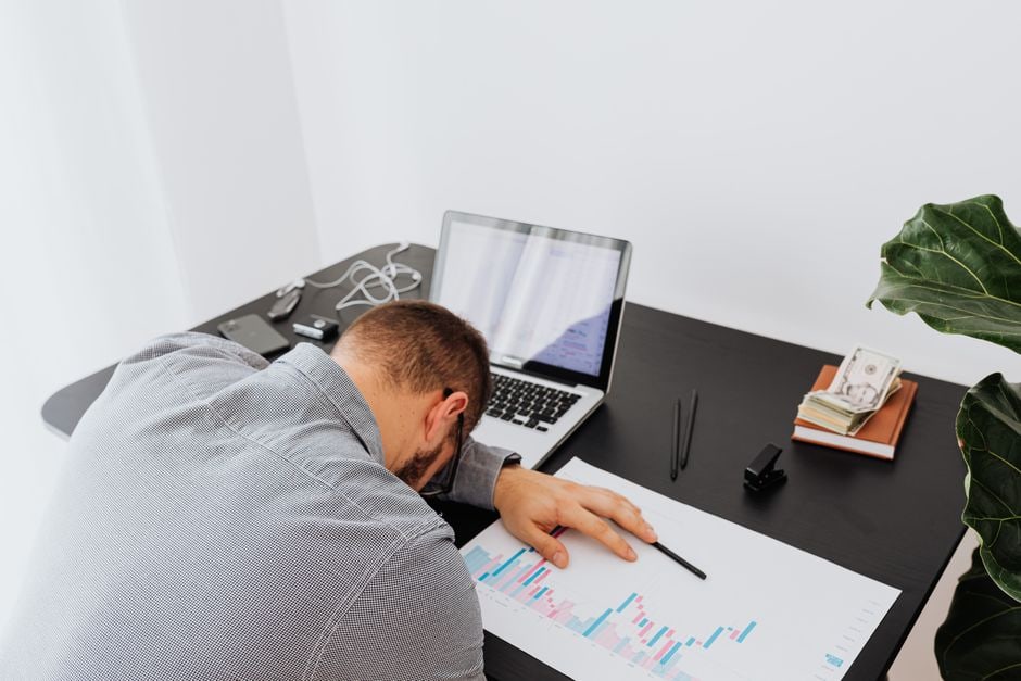A busy professional sits at a laptop in a modern office, head in hands, looking visibly exhausted and stressed, with papers scattered around, illustrating the need for rest.