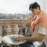 A person works on a laptop on a balcony overlooking a bustling European city, symbolizing the dream of a workcation.