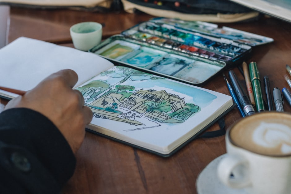 A remote worker sips coffee while sketching in a notebook, taking a creative break from work.