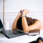 A remote worker hunched over a laptop at a home desk, surrounded by papers, looking overwhelmed and stressed.