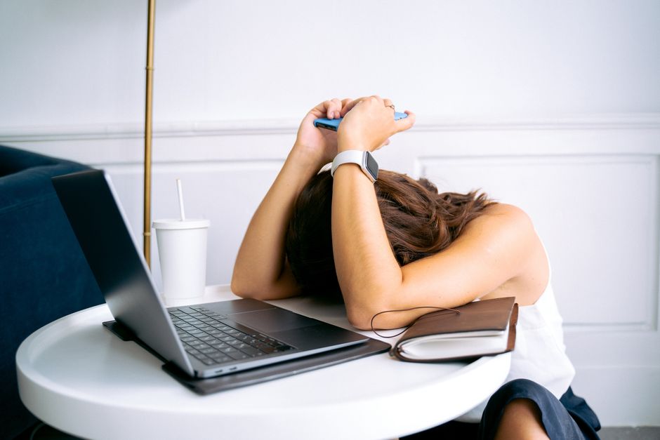 A remote worker hunched over a laptop at a home desk, surrounded by papers, looking overwhelmed and stressed.