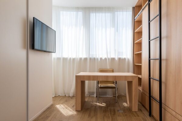 A neatly organized small home office setup in a cozy apartment, featuring a wall-mounted monitor and floating shelves maximizing vertical space.