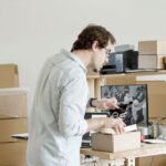 A small business owner looking thoughtfully at different printer boxes in a store aisle, weighing options for occasional use.