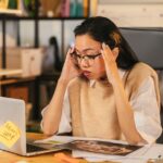 A small business owner sits at a desk, looking stressed while scrolling on a laptop, surrounded by paperwork and an unfinished coffee, symbolizing the common challenges of managing customer inquiries.