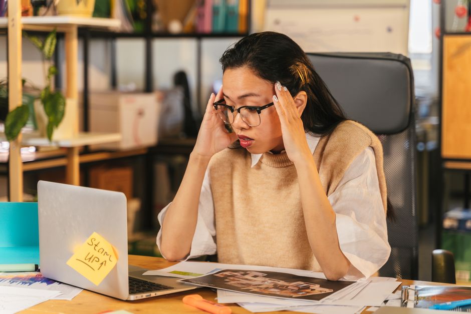 A small business owner sits at a desk, looking stressed while scrolling on a laptop, surrounded by paperwork and an unfinished coffee, symbolizing the common challenges of managing customer inquiries.