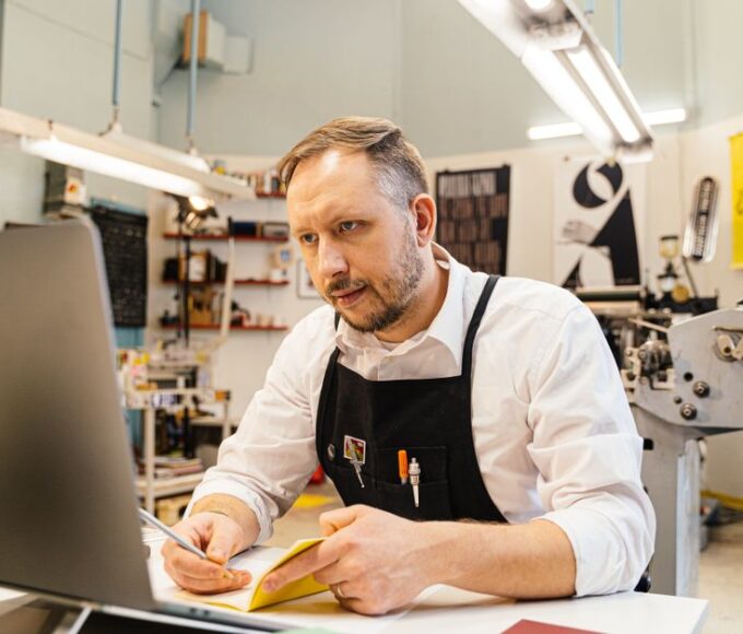 A small business owner smiling while typing on a laptop, with subtle digital overlays suggesting AI assistance, in a well-lit cafe setting.