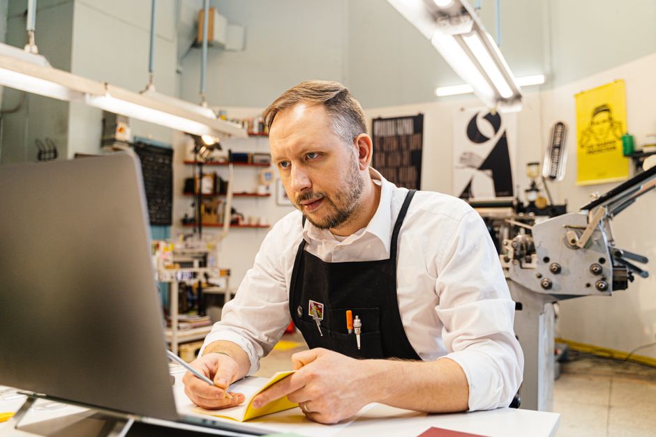 A small business owner smiling while typing on a laptop, with subtle digital overlays suggesting AI assistance, in a well-lit cafe setting.